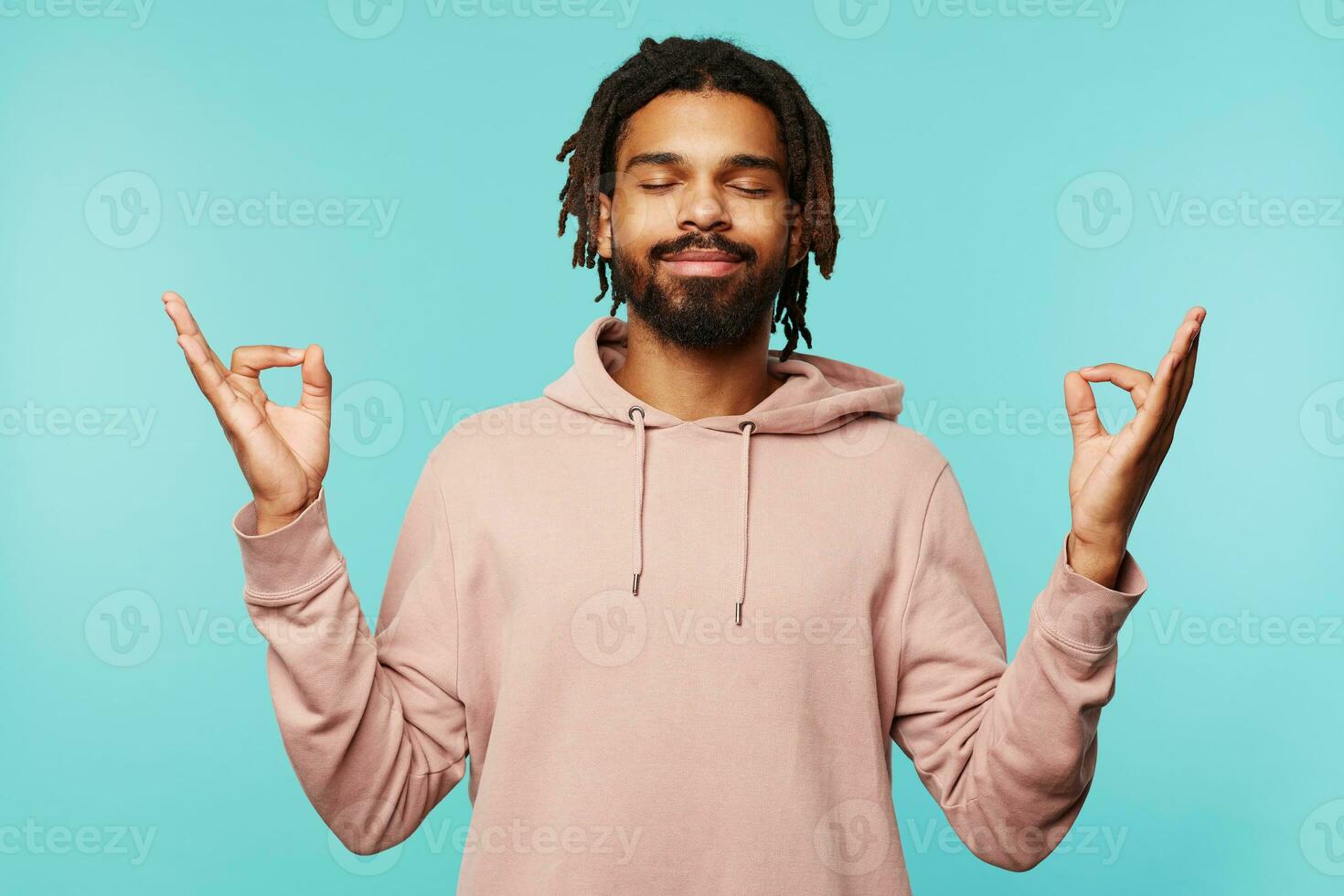 Good looking young dark skinned bearded guy with dreadlocks keeping his eyes closed while smiling pleasantly, posing over blue background with raised hands photo