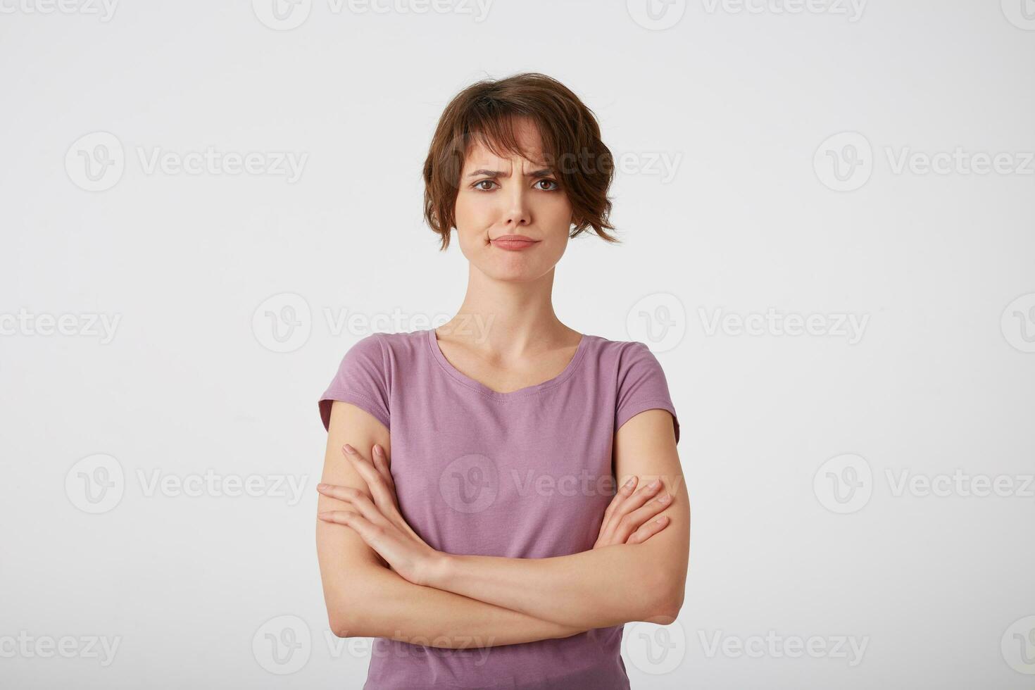 Portrait of frowning displeased short-haired lady in blank t-shirt, doubts the decision with arms crossed, stands over white wall. photo