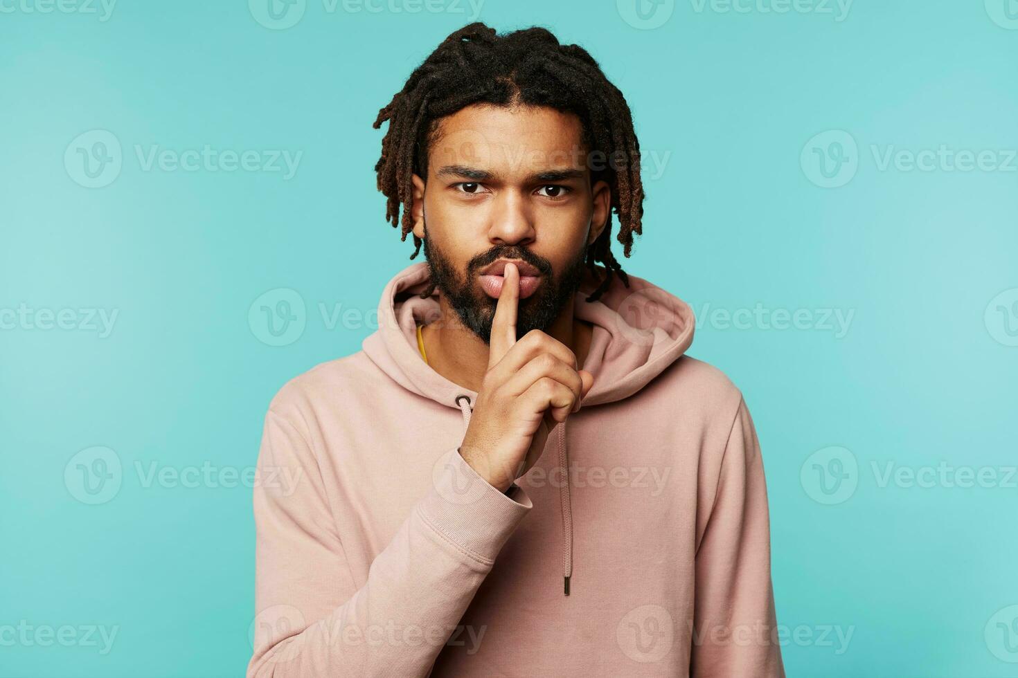 Serious young brown-eyed dark haired bearded man raising hand with hush gesture to his mouth while asking to keep silence, standing over blue background photo