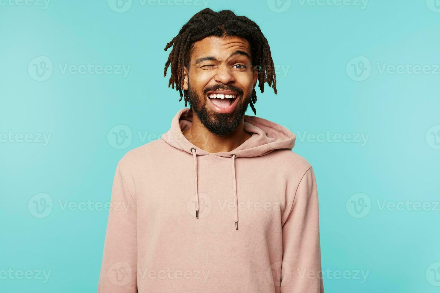 Joyful young pretty brunette male with dark skin winking happily at camera and smiling cheerfully, keeping hands down while standing over blue background photo