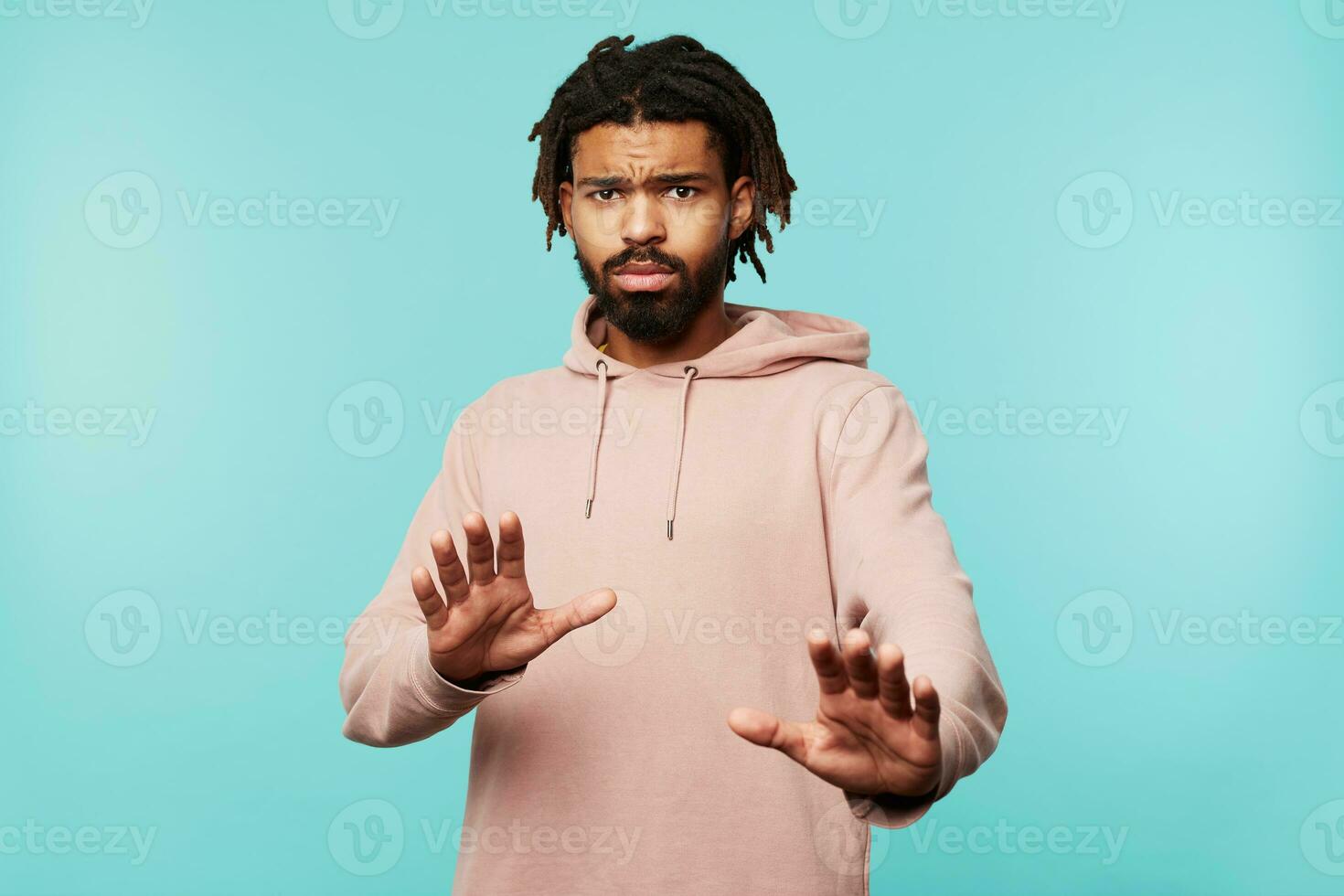 Frightened young dark skinned bearded male with dreadlocks raising hands in protective gesture and looking scaredly at camera, posing over blue background photo