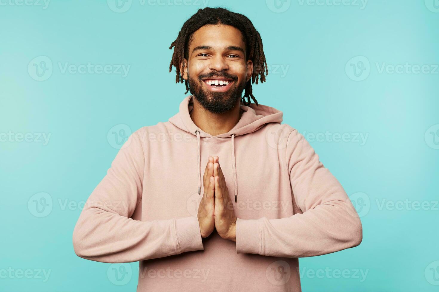 Cheerful young attractive brown haired guy with beard smiling widely at camera and folding raised palms together, dressed in pink hoodie while posing over blue background photo