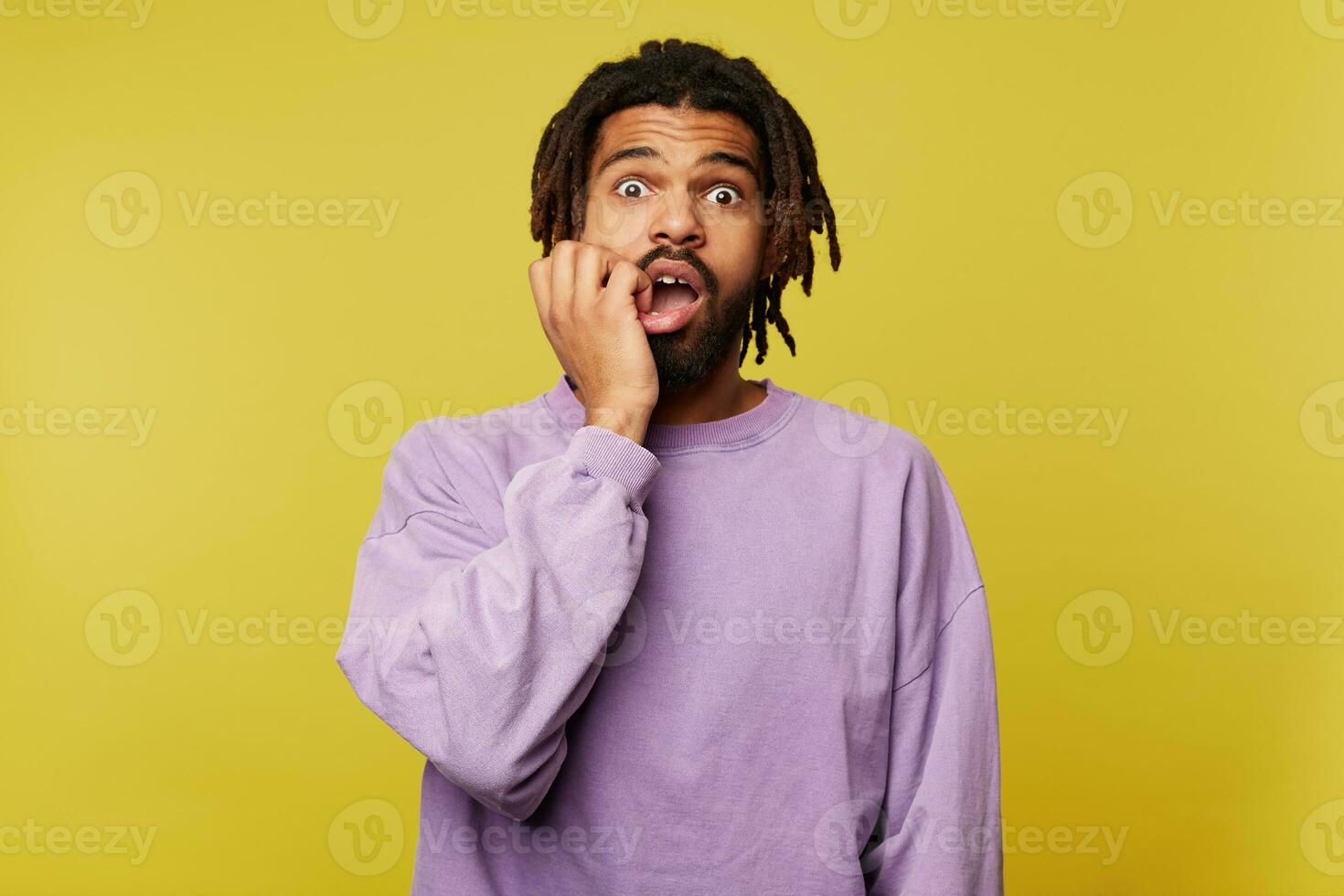 Afraid young attractive brown haired bearded man with dark skin looking at camera with wide eyes opened and holding scaredly hand on his face, isolated over yellow background photo