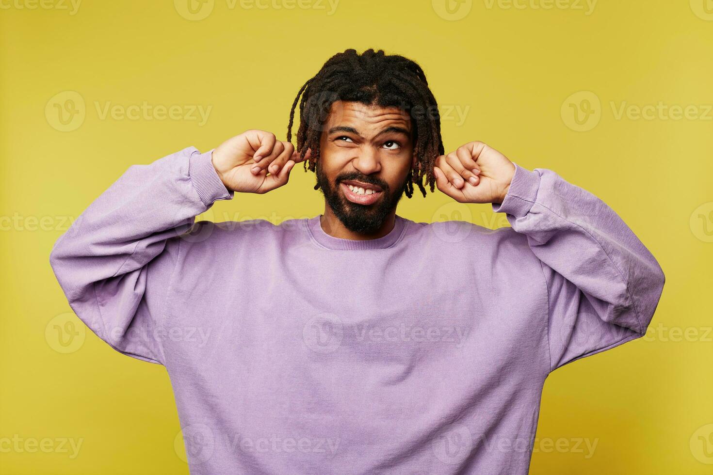 Displeased young dark skinned brunette bearded man with dreadlocks frowning his face while looking confusedly upwards and inserting forefingers into his ears, isolated over yellow background photo