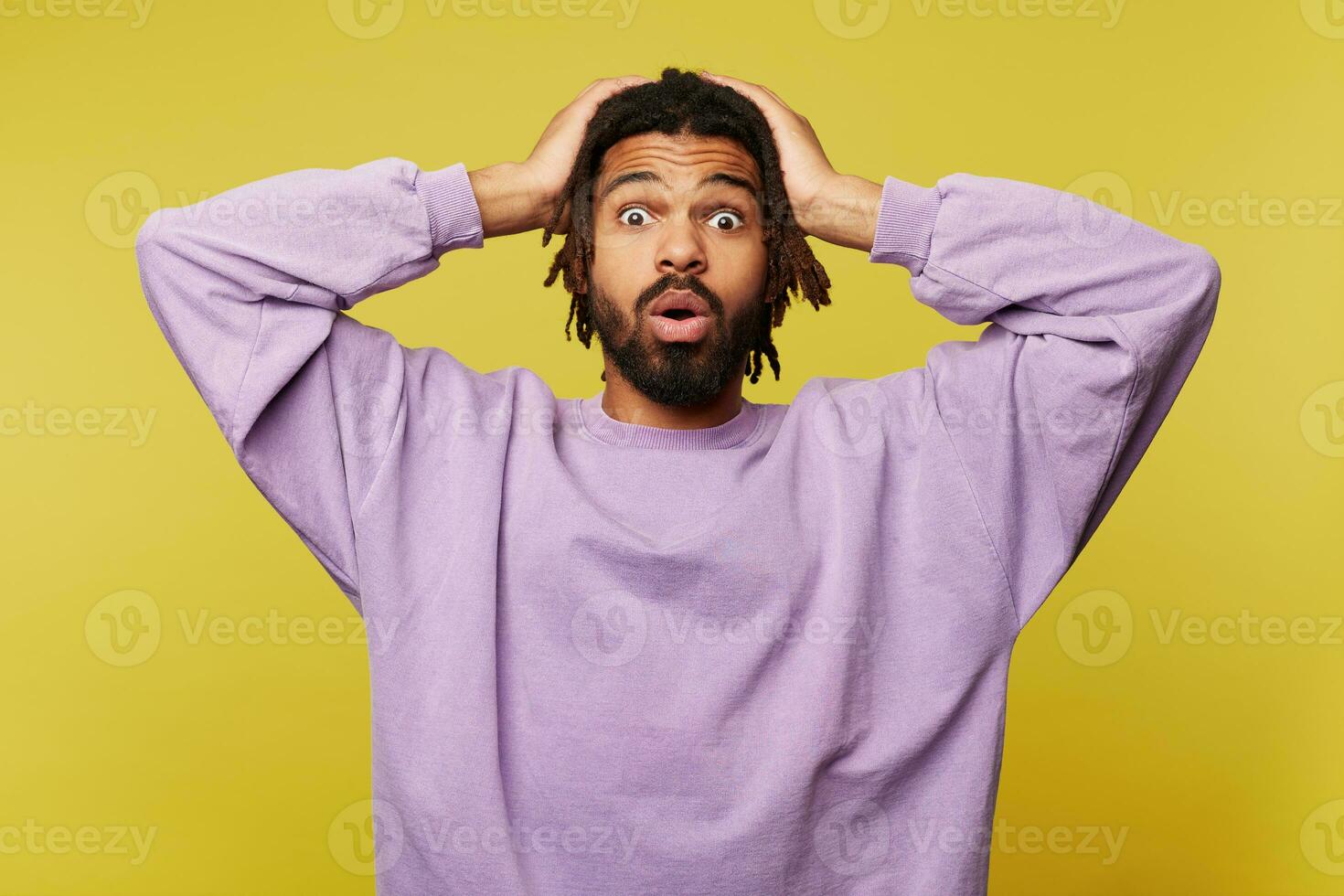 Shocked young handosme dark skinned bearded man clutching his head with raised hands and looking amazedly at camera, isolated against yellow background photo