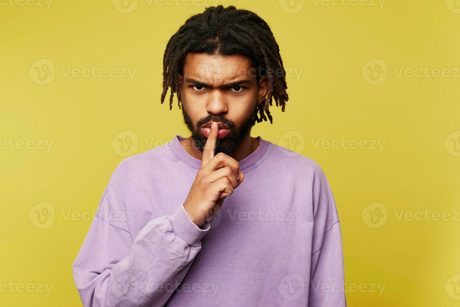 Severe young handsome dark skinned brunette bearded guy with dreadlocks frowning his eyebrows while looking seriously at camera and holding forefinger on his lips, isolated over yellow background photo