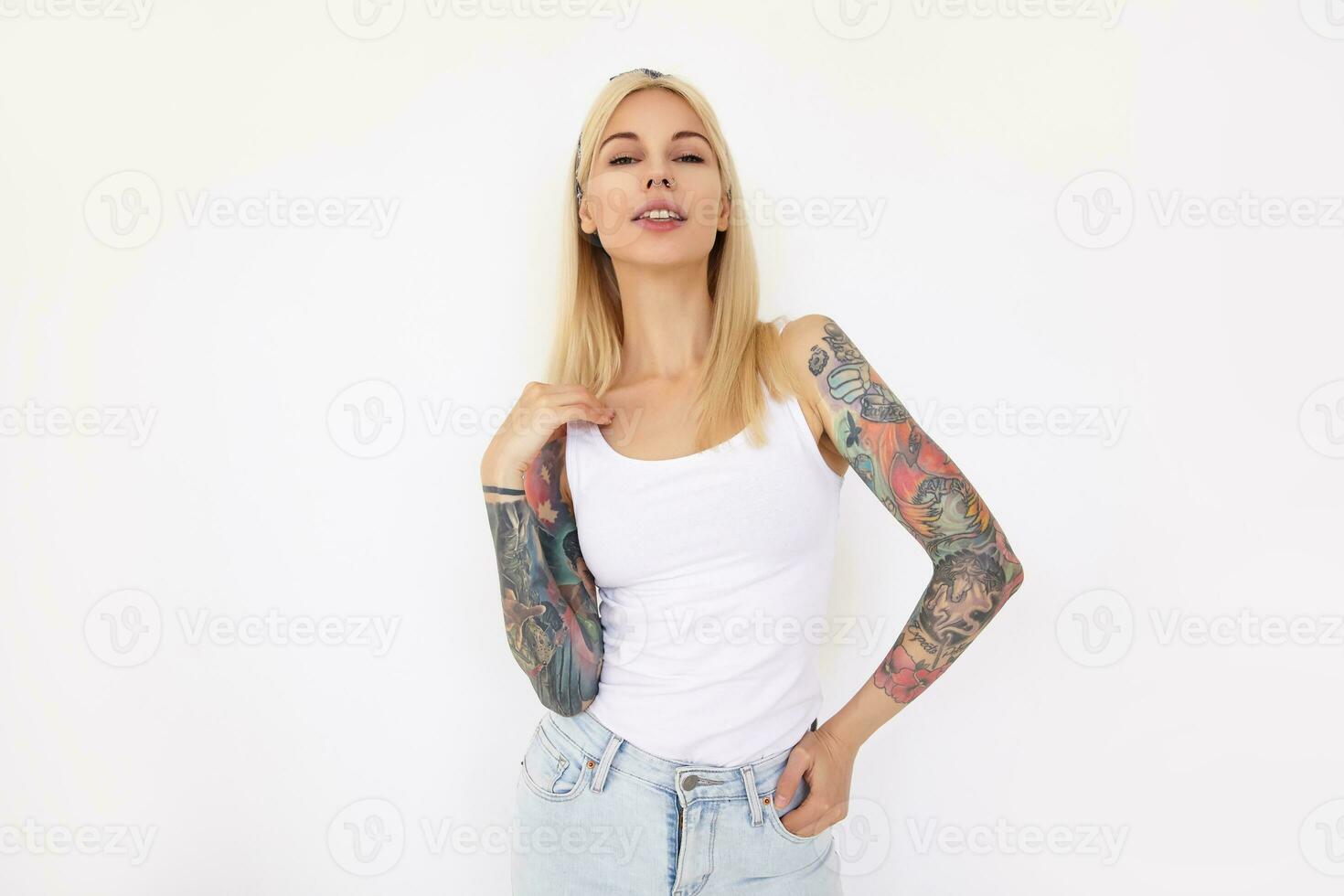 Beautiful young tattooed blonde woman with casual hairstyle raising hand to her hair while looking at camera, isolated over white background in headband and casual wear photo