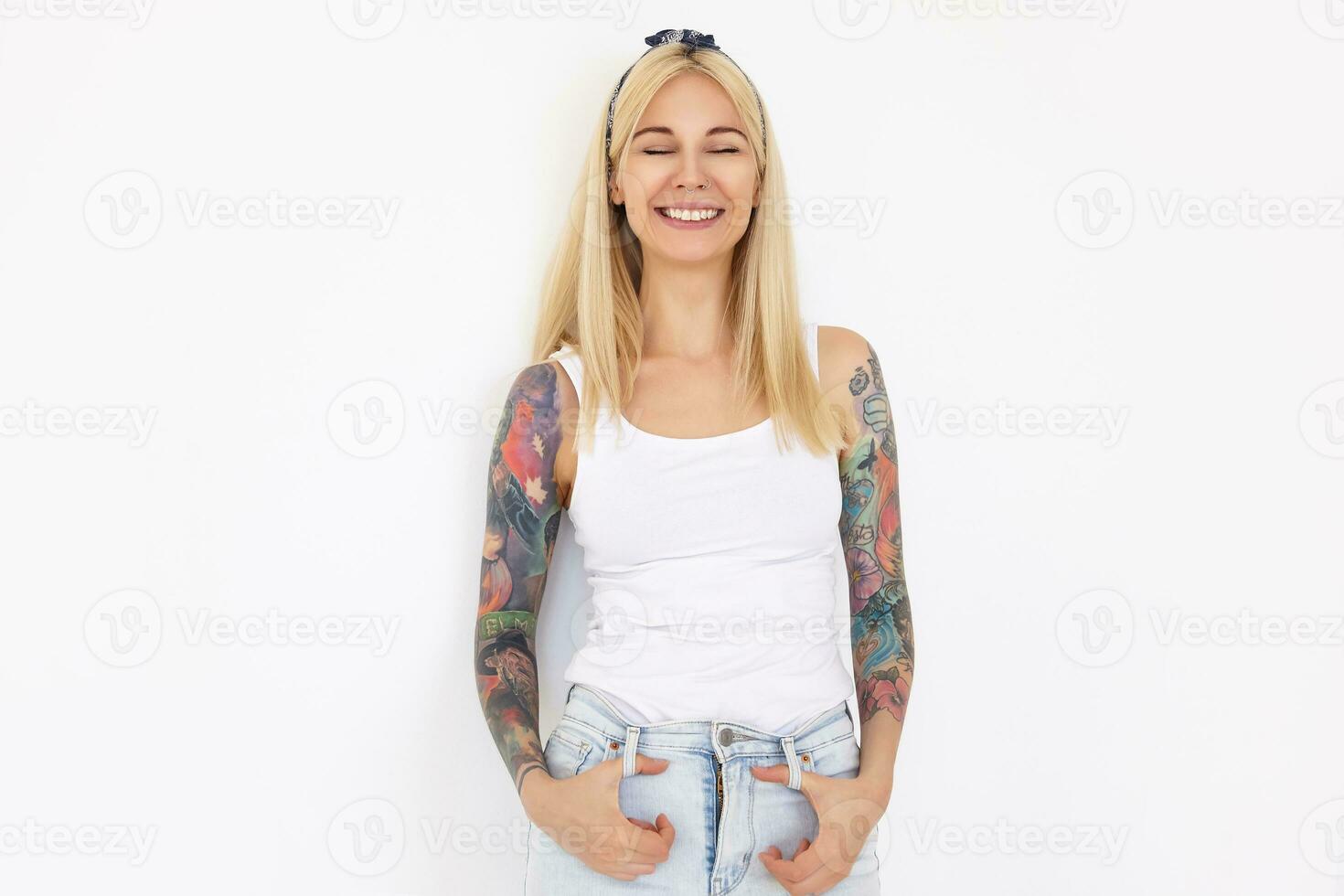 Good looking young happy long haired lady with tattoos wearing headband and casual clothes while posing over white background, keeping her eyes closed while smiling gladly photo