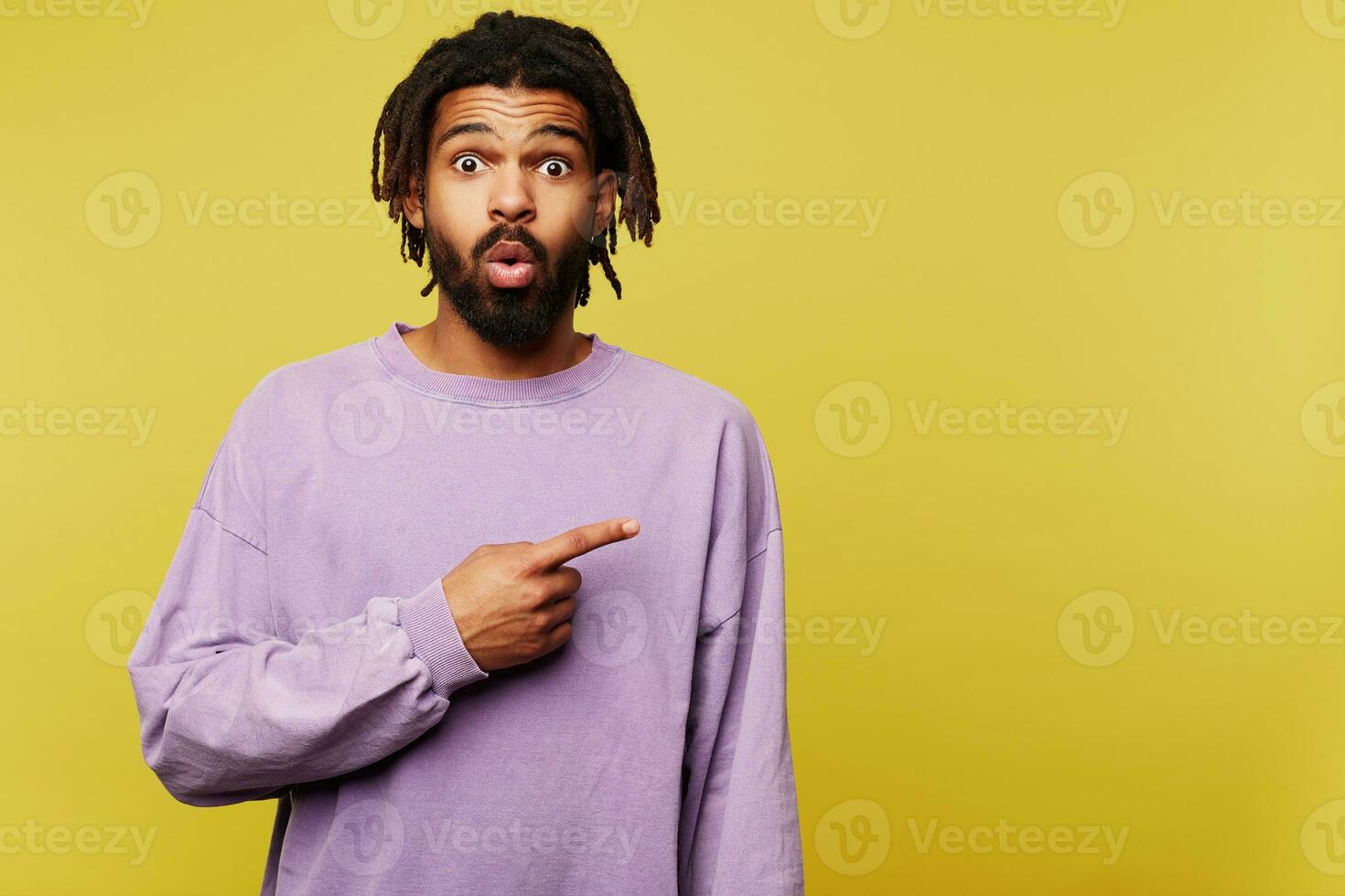 Open-eyed young attractive dark skinned brunette bearded male showing aside with forefinger and raising surprisedly eyebrows, standing against yellow background photo