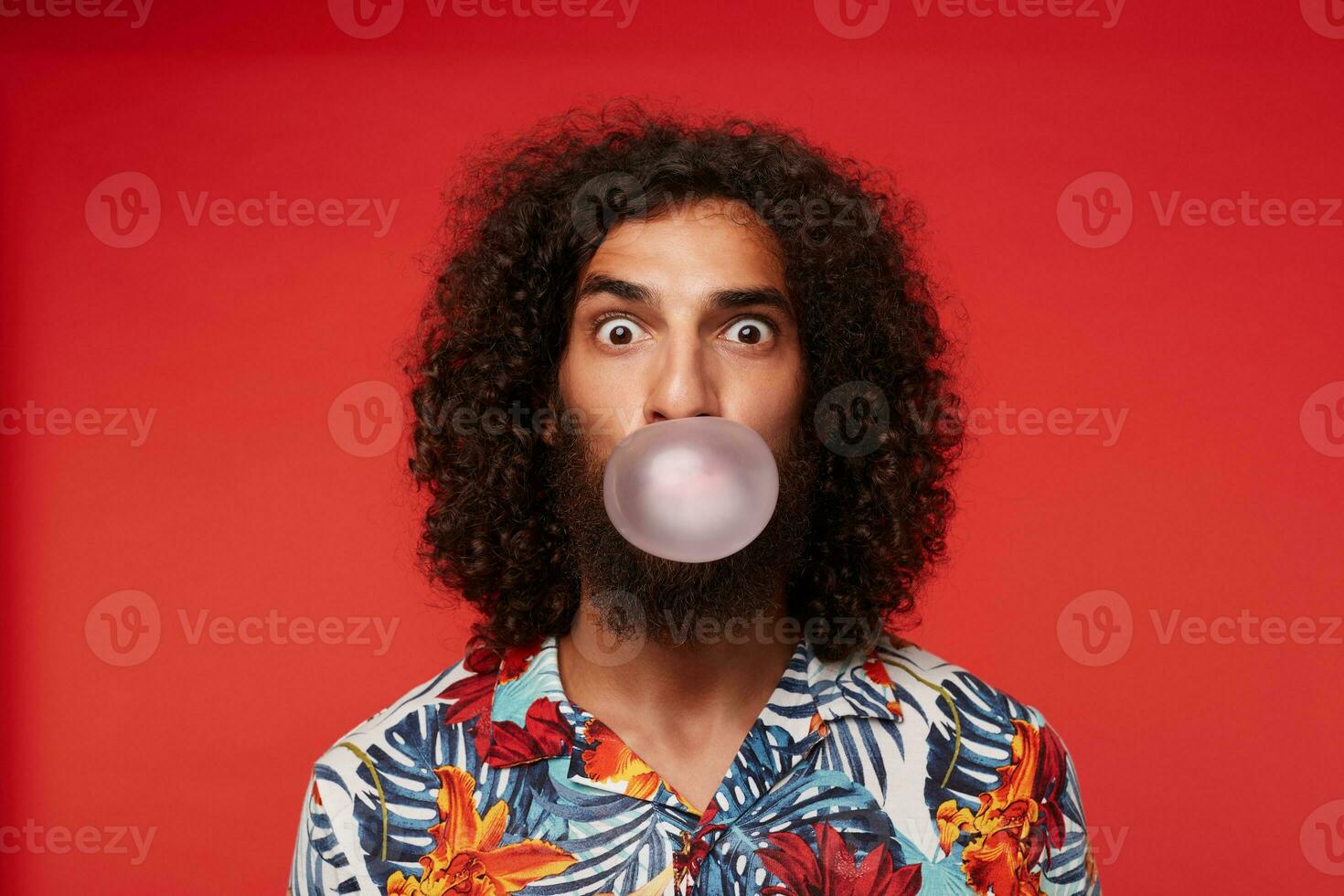 Amazed pretty brown-eyed bearded guy with dark curly hair making bubble with gum and looking surprisedly to camera, standing over red background in shirt with floral print photo