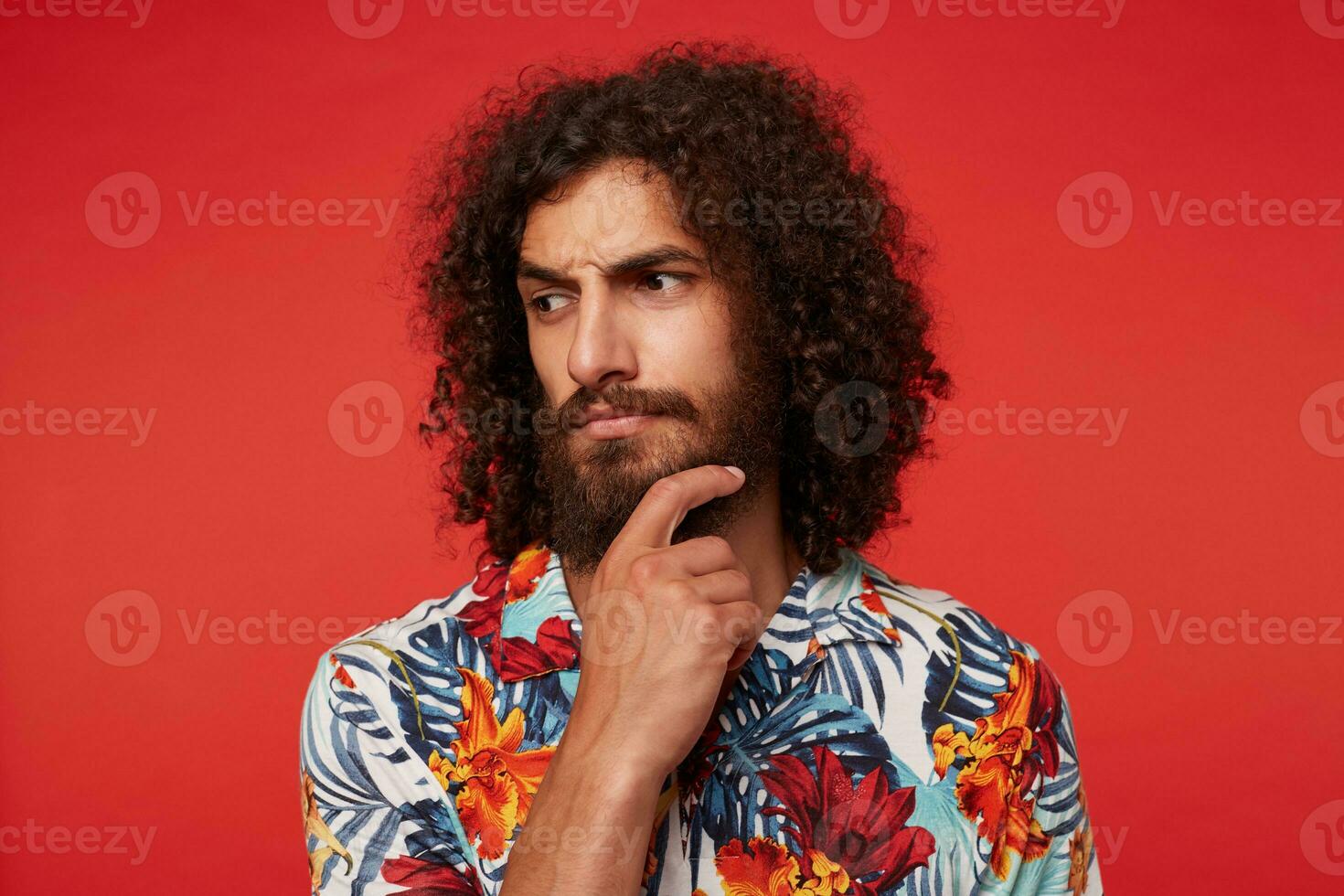 Studio photo of pensive young brunette curly male with lush beard looking thoughtfully aside and frowning eyebrows, keeping raised hand on his chin while posing against red background