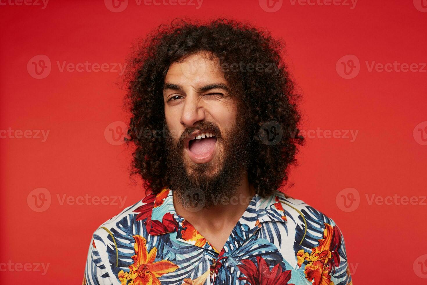 Close-up of cheerful young brunette curly man with beard winking at camera and keeping mouth wide opened, dressed in shirt with floral print against red background photo