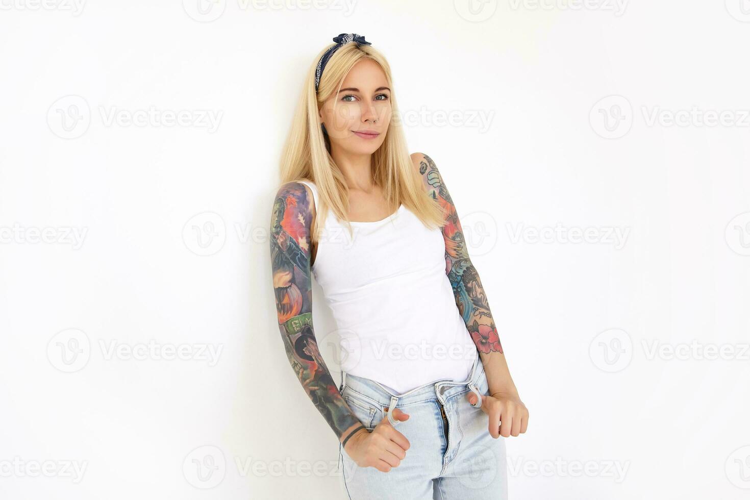 Glad young beautiful tattooed blonde lady with casual hairstyle looking at camera with pleasant smile while standing over white background and keeping hands on her jeans photo