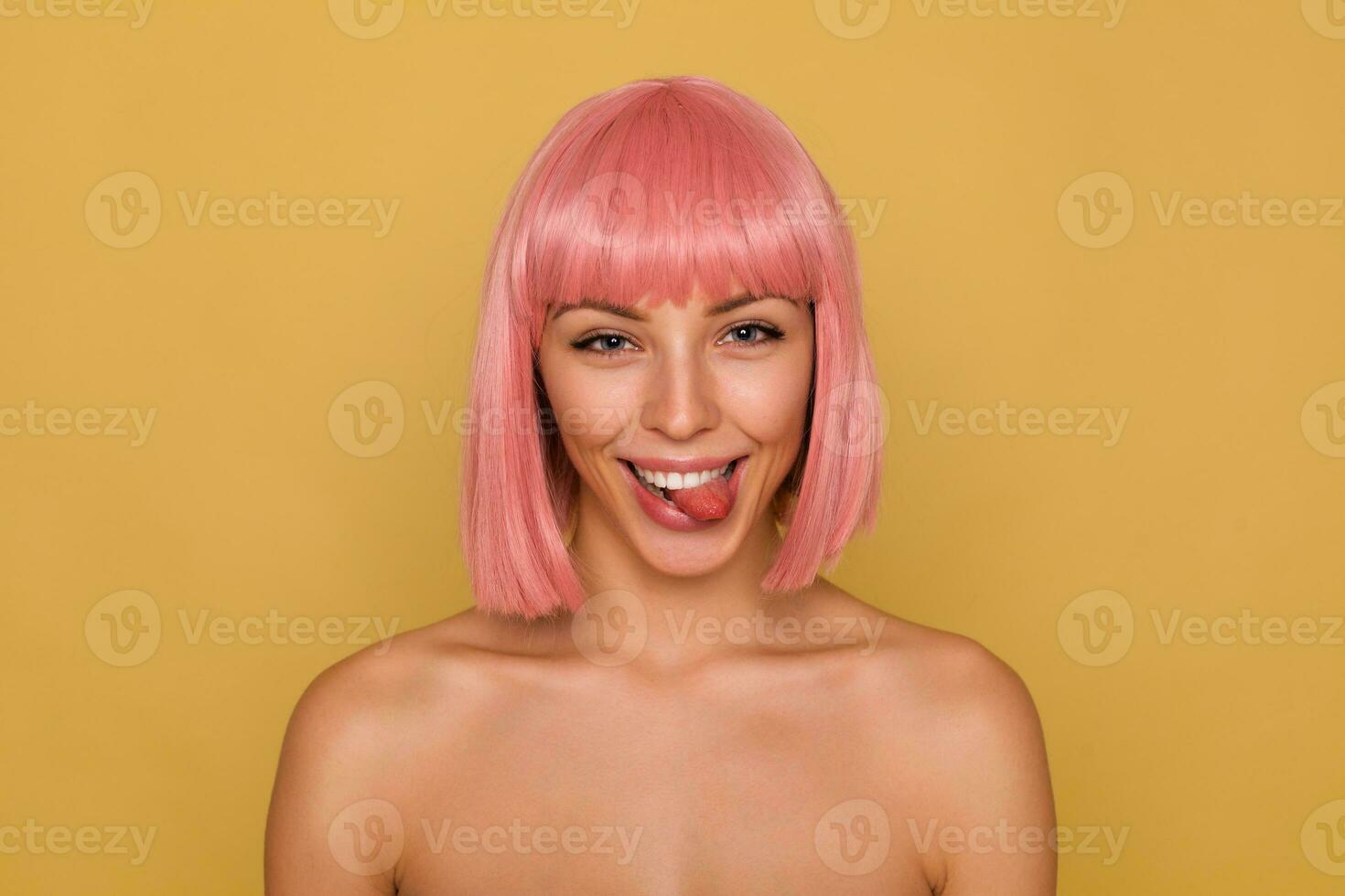 Funny shot of young pretty joyful lady with short pink hair looking playfully at camera and showing her tongue, fooling while posing over mustard background photo