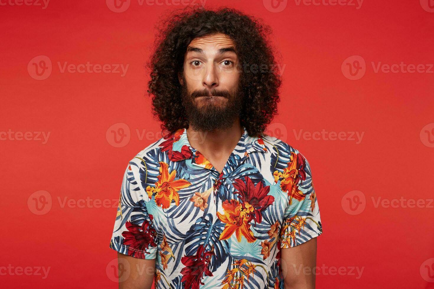 Surprised attractive brunette curly guy with beard biting his lips and looking to camera with raised eyebrows, standing over red background with hands along body photo