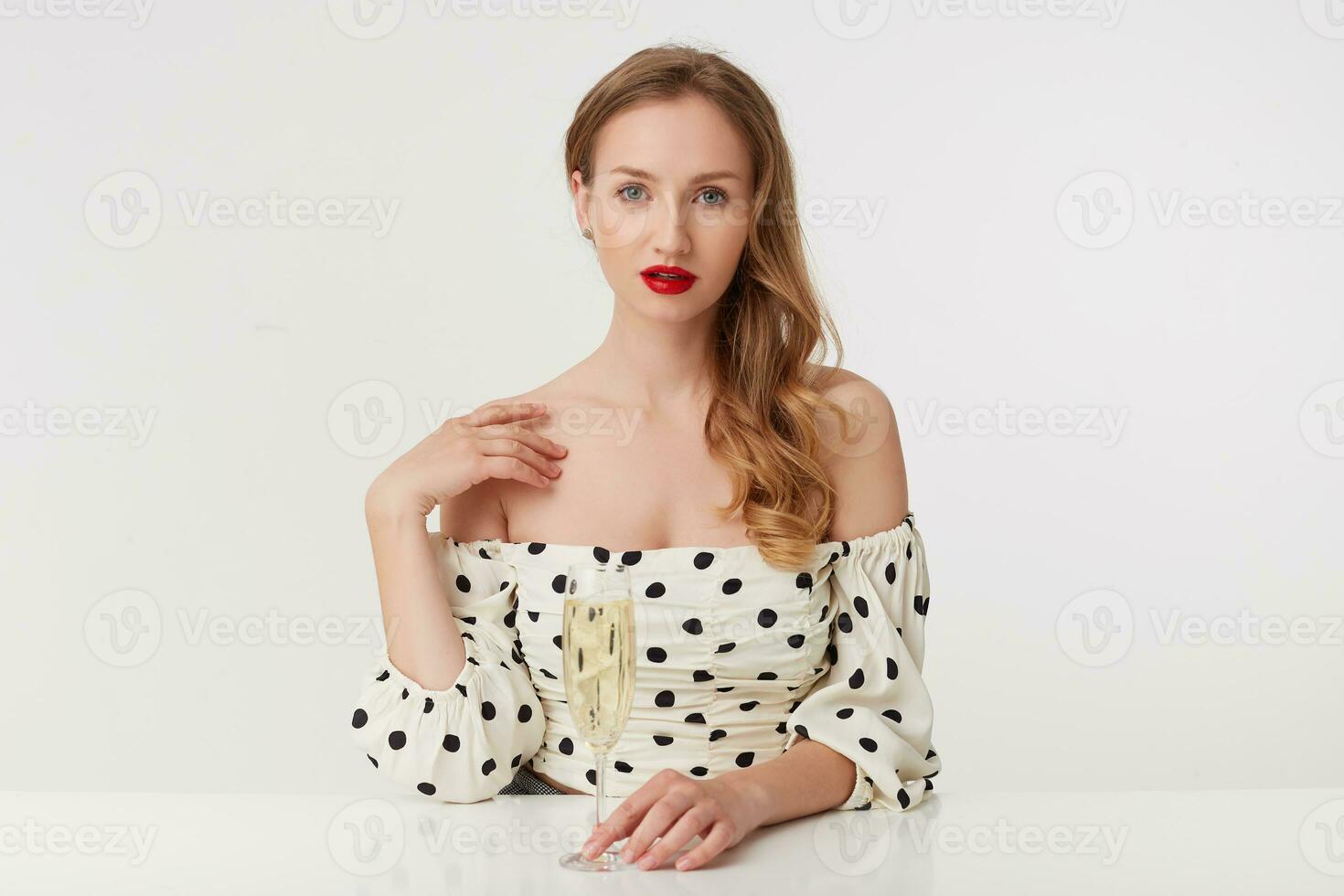 blue-eyed girl with long blond hair, with red lips in a polka-dot dress, sits at the table, puts her hand to her shoulder and holds a glass of champagne. Isolated over white white background. photo