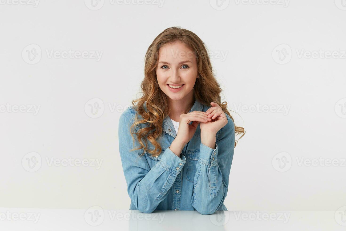 estudio Disparo de sonriente joven linda mujer sentado a el mesa, con largo rubio ondulado cabello, vistiendo un mezclilla camisa. mirando a el cámara aislado terminado blanco antecedentes. foto