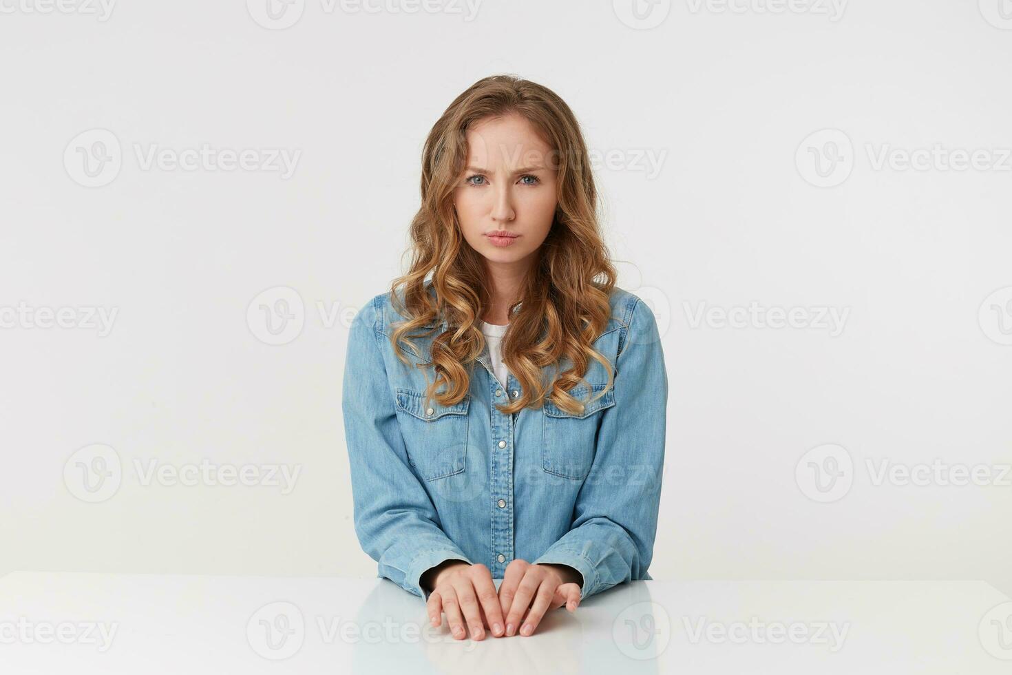 Studio shot of displeased young beautiful woman with long blond wavy hair, wearing a denim shirt, siting at the table, frowning and looking at the camera isolated over white background. photo