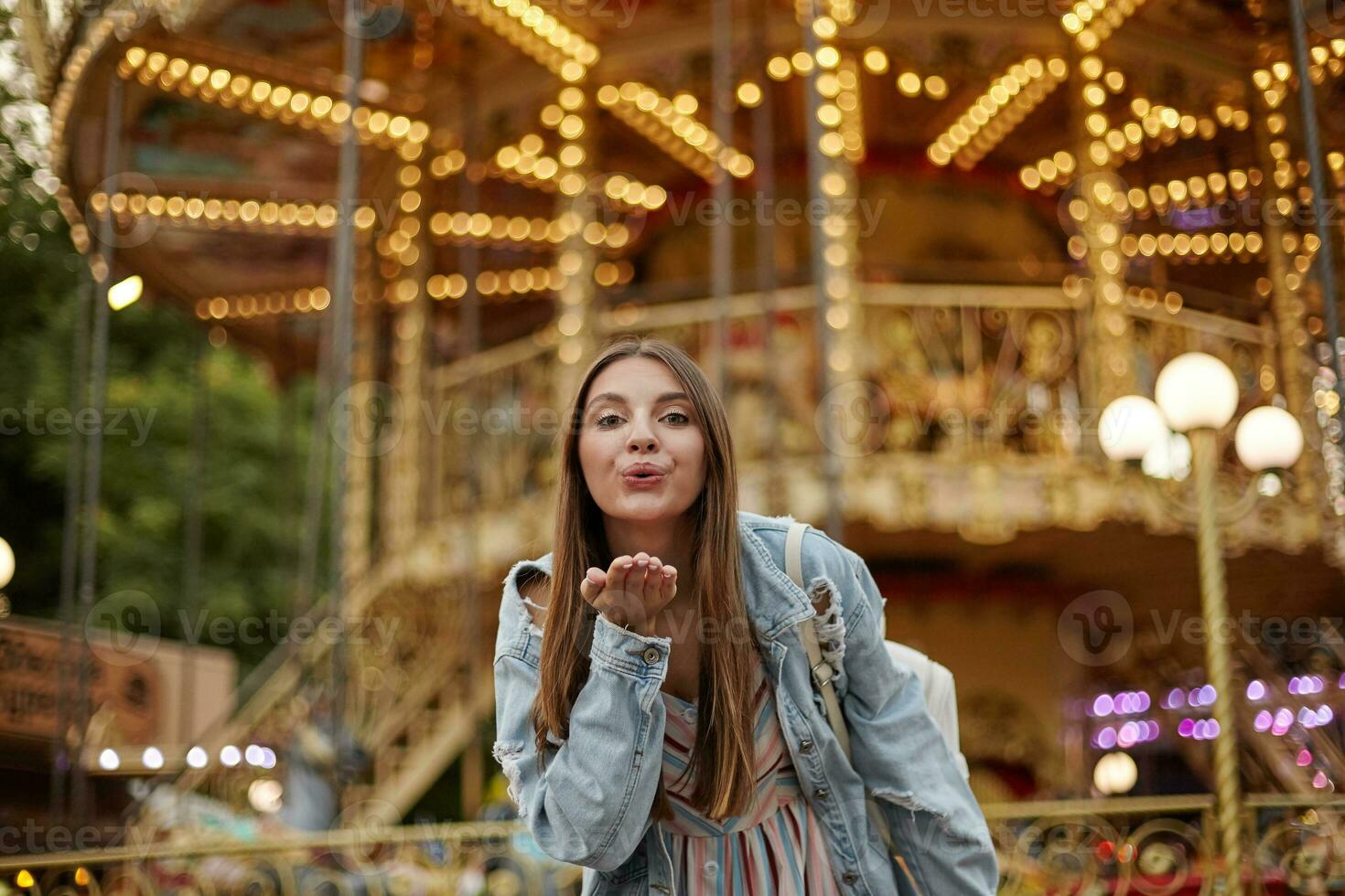 Outdoor photo of charming long haired young lady in trendy jeans coat raising palm and blowing air kiss to camera, standing over carousel in park of attractions