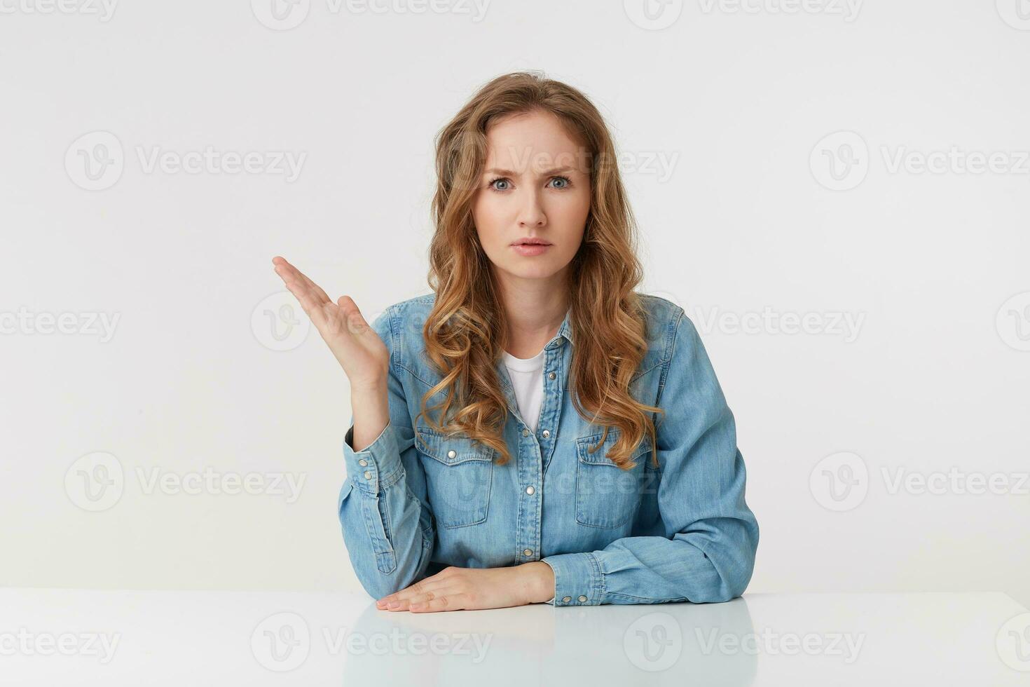 Portrait of at a loss young woman with long blond wavy hair, sitting at the table, one palm raised, looks skeptically displeased indignantly, isolated over white background. photo