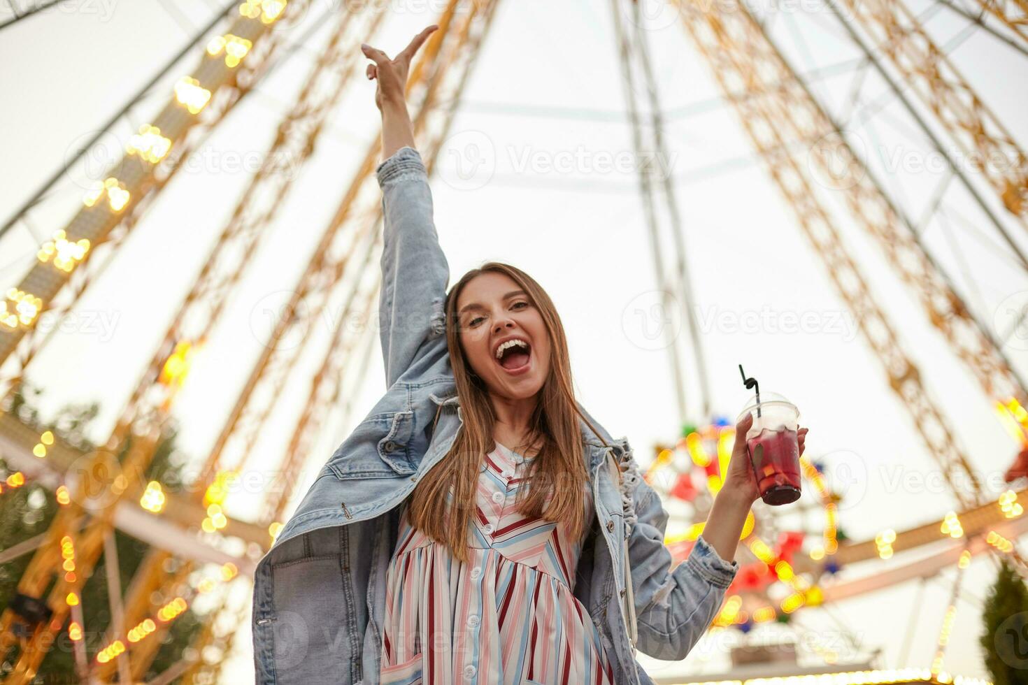 Positive young beautiful brunette woman posing over attractions in amusement park with cup of lemonade in hand, raising palm upwards and smiling broadly to camera photo