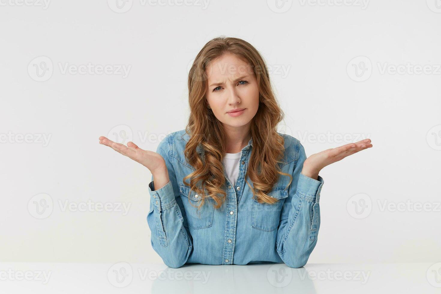 Confused young woman with long blond wavy hair, wearing a denim shirt, throws up his hands, frowning and looking at the camera isolated over white background. photo