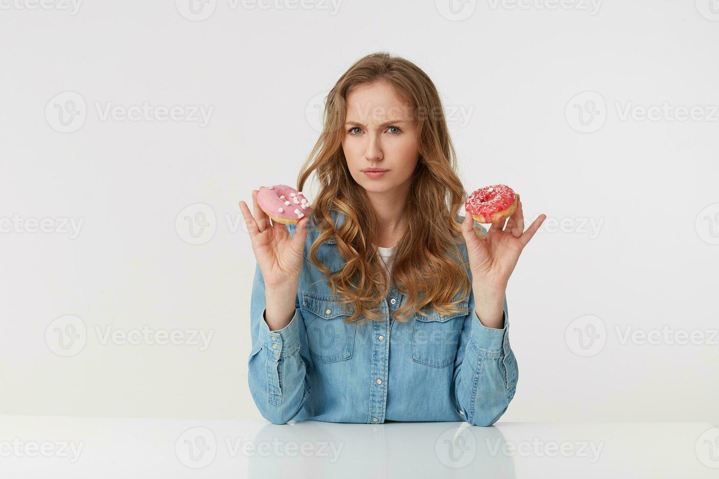 Young beautiful girl holds donuts in her hands, wearing a denim shirt, promotes healthy eating and give up sugar. Looks disapproving of the camera isolated over white background. photo