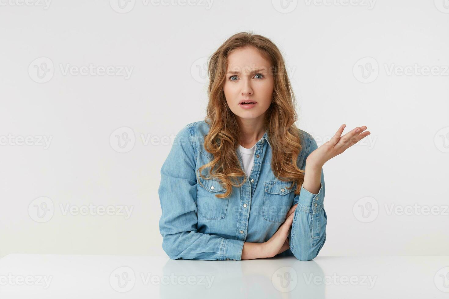 Portrait of confused young woman with long blond wavy hair, sitting at the table, one palm raised, looks skeptically displeased indignantly with incomprehension, over white background. photo