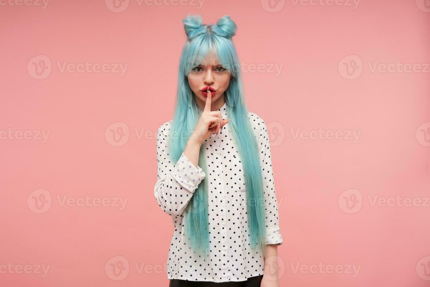 Studio photo of lovely young lady with long blue hair keeping forefinger on her lips while looking seriously at camera, asking to keep silence while posing over pink background