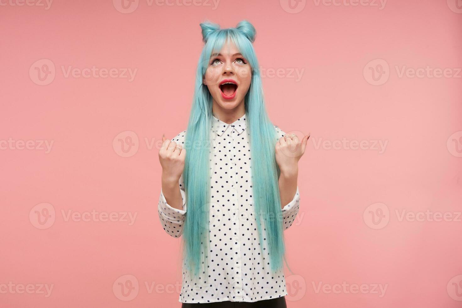 Surprised young attractive long haired woman with bright makeup looking excitedly upwards with wide mouth opened, dressed in white shirt with black dots over pink background photo