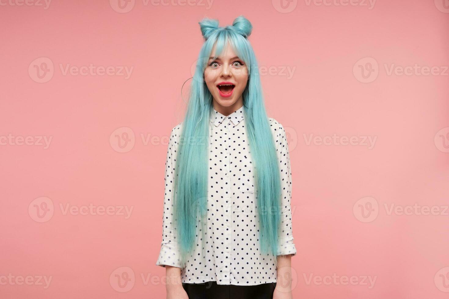 Open-eyed young attractive lady with long blue hair looking amazedly at camera and keeping mouth wide opened, dressed in shirt with black dots while standing over pink background photo