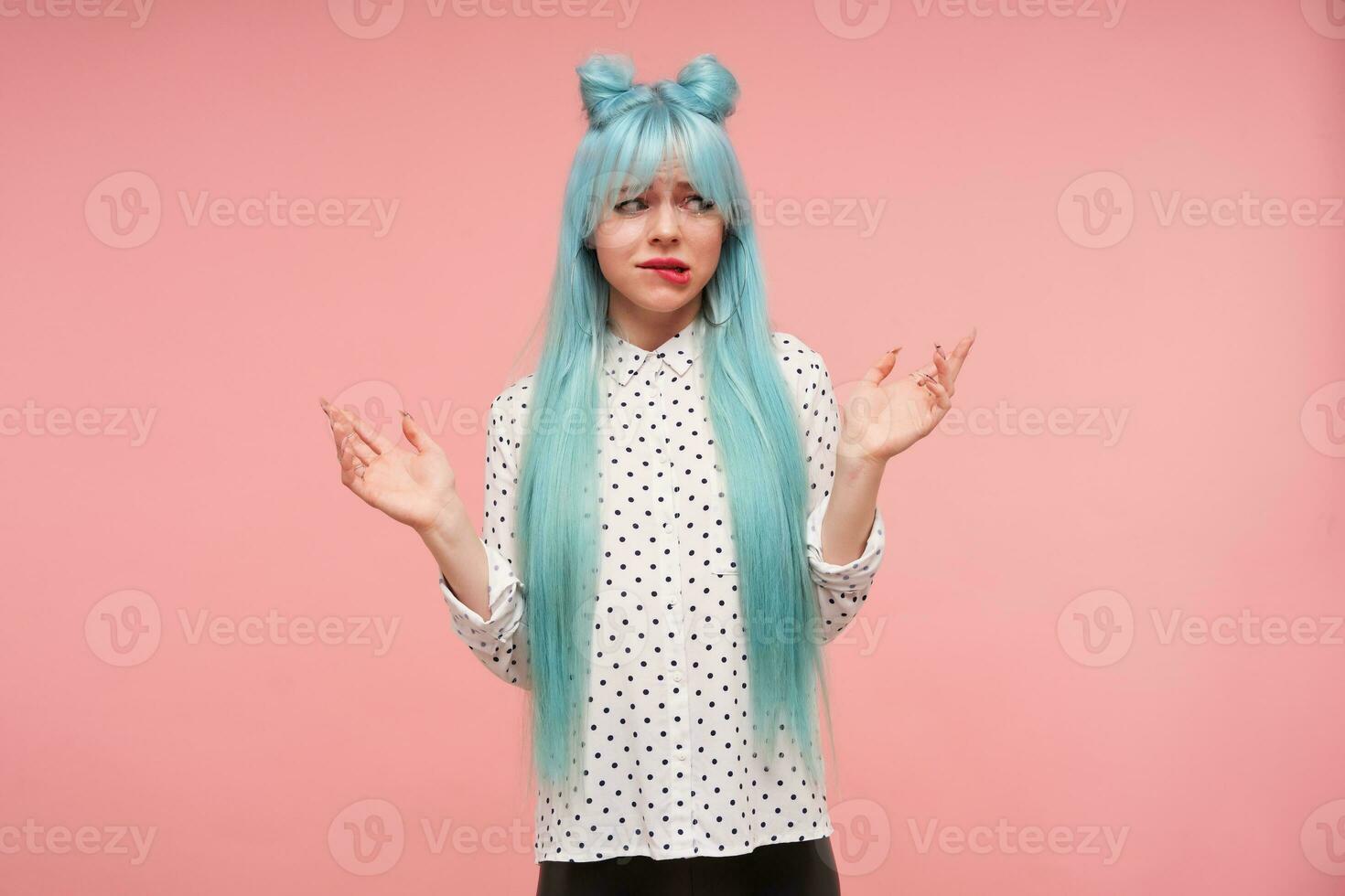 Bewildered attractive young blue haired woman frowning her eyebrows and biting discomposedly underlip, keeping palms raised while standing over pink background photo