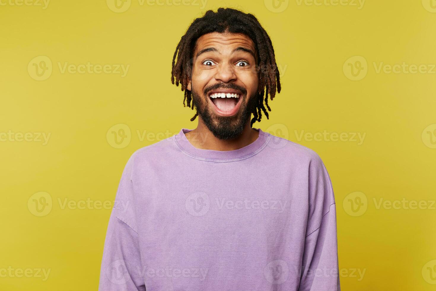 Joyful young attractive dark skinned brown haired man with dreadlocks looking surprisedly at camera with opened mouth while standing over yellow background photo