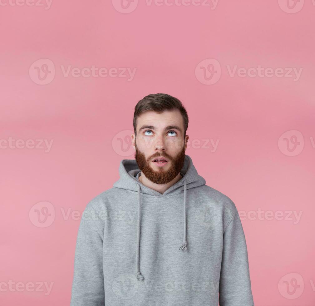 Portrait of young tired handsome guy looking up at the copy space, thinking about solving a problem, presents possible options, stands over pink background. photo
