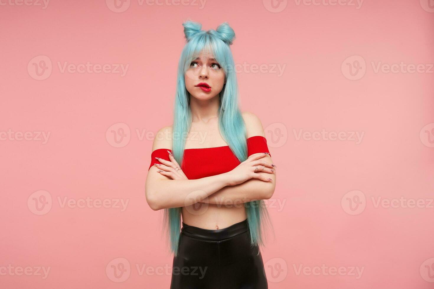 Indoor shot of beautiful young woman with long blue anime hair crossing hands on her chest and looking thoughtfully upwards,biting underlip while posing over pink background in red top and black pants photo