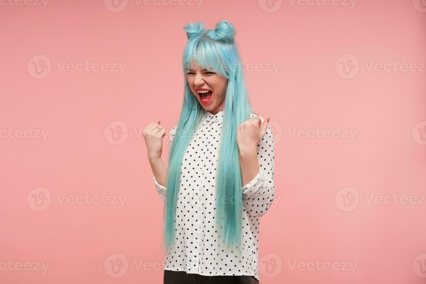 Studio photo of excited pretty young blue haired female raising happily fists and shouting joyfully, rejoicing about something nice happened, standing over pink background