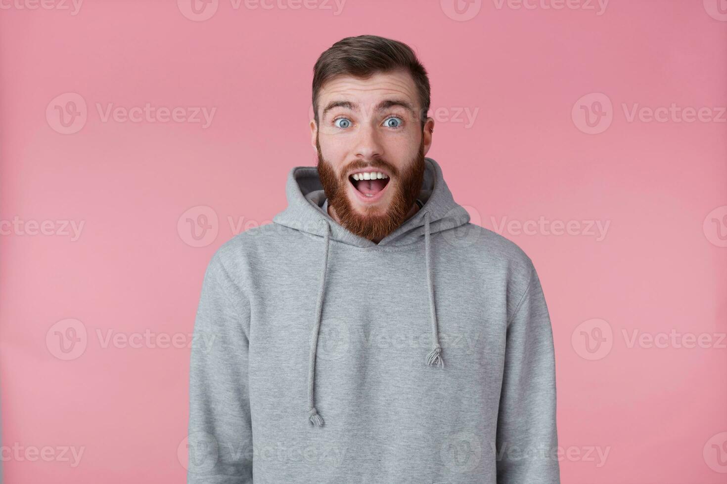 Photo of young handsome happy amazed red bearded man in gray hoodie, looks surprised, stands over pink background with wide open mouth and eyes.