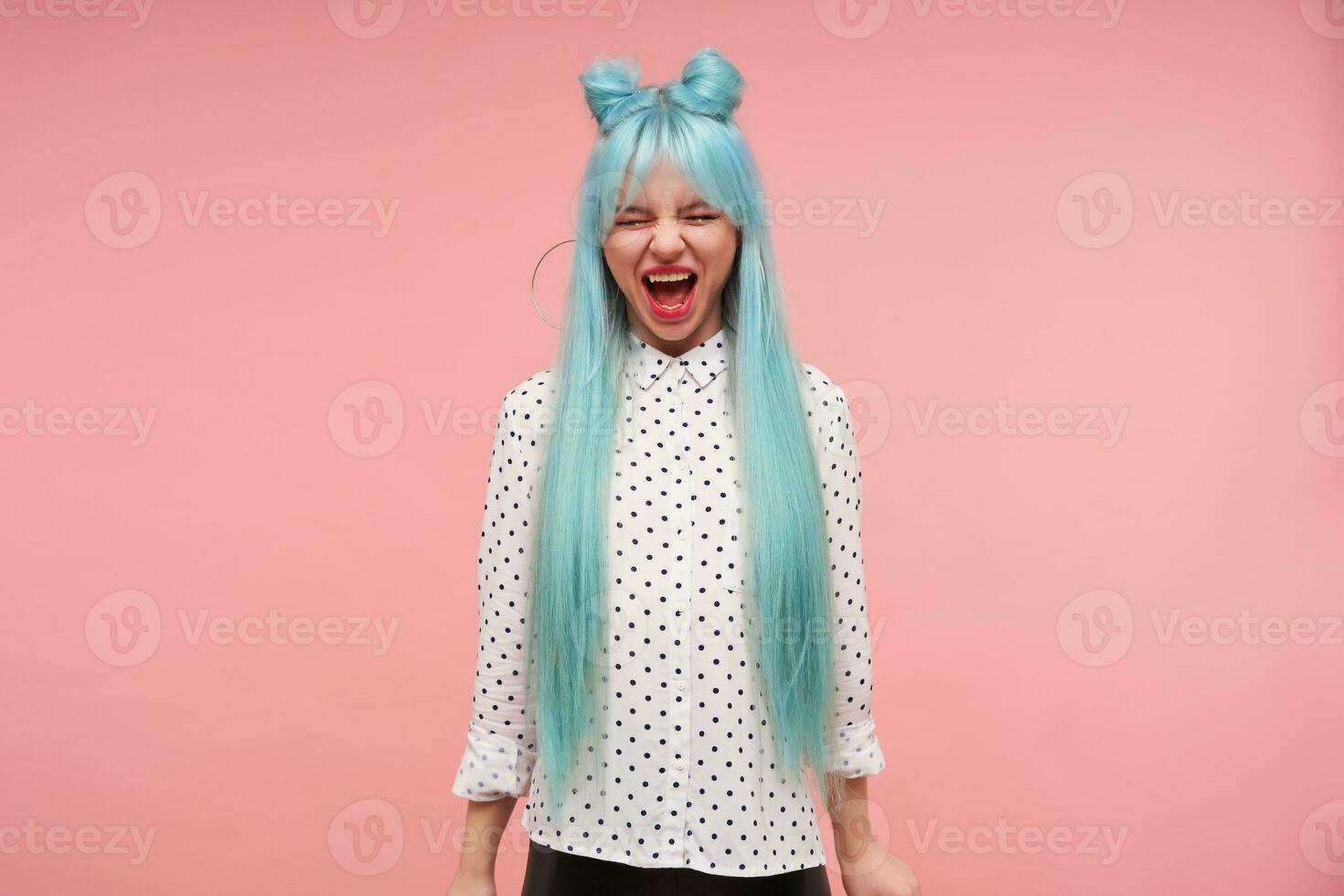 Indoor shot of young lovely female with blue anime hair keeping her eyes closed while screaming loud, wearing buns hairstyle while standing over pink background with hands down photo