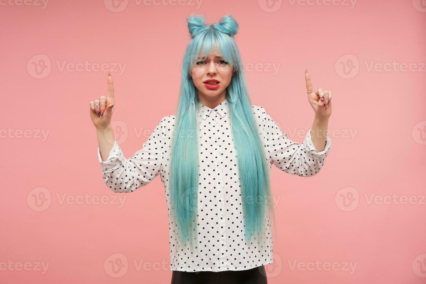 disgustado joven azul peludo dama ceñudo su cara y mirando a cámara con abadejo, levantamiento dedos indices y señalando hacia arriba, vestido en blanco camisa con negro puntos terminado rosado antecedentes foto