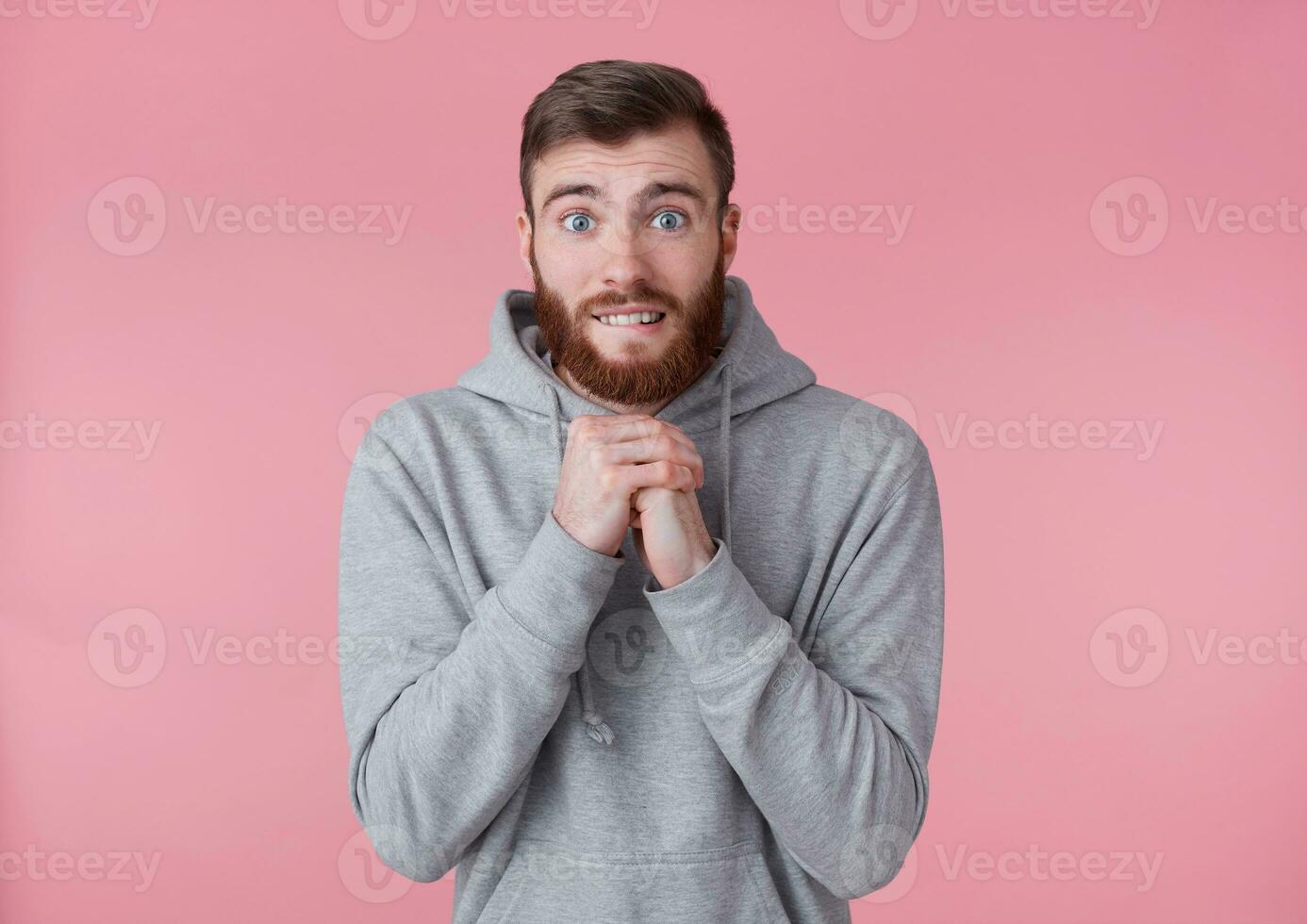 Portrait of young attractive red bearded man in gray hoodie, looks at the camera, cross palms and asking for something, hope for good luck. Stands over pink background. photo