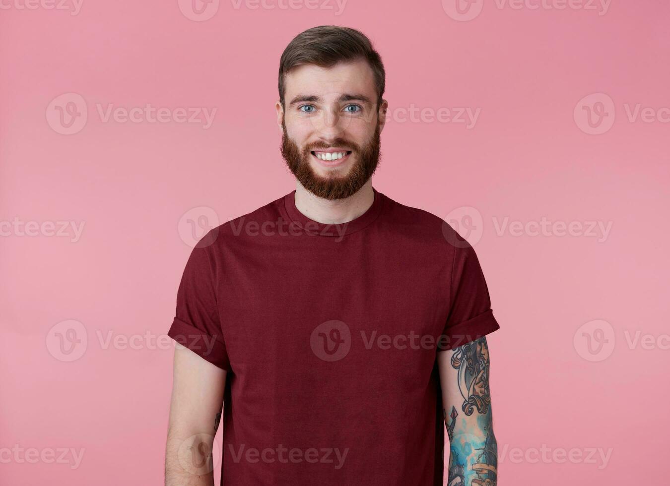 Photo of young handsome cheerful red bearded man in red t-shirt, looks happy and broadly smiles, stands over pink background.