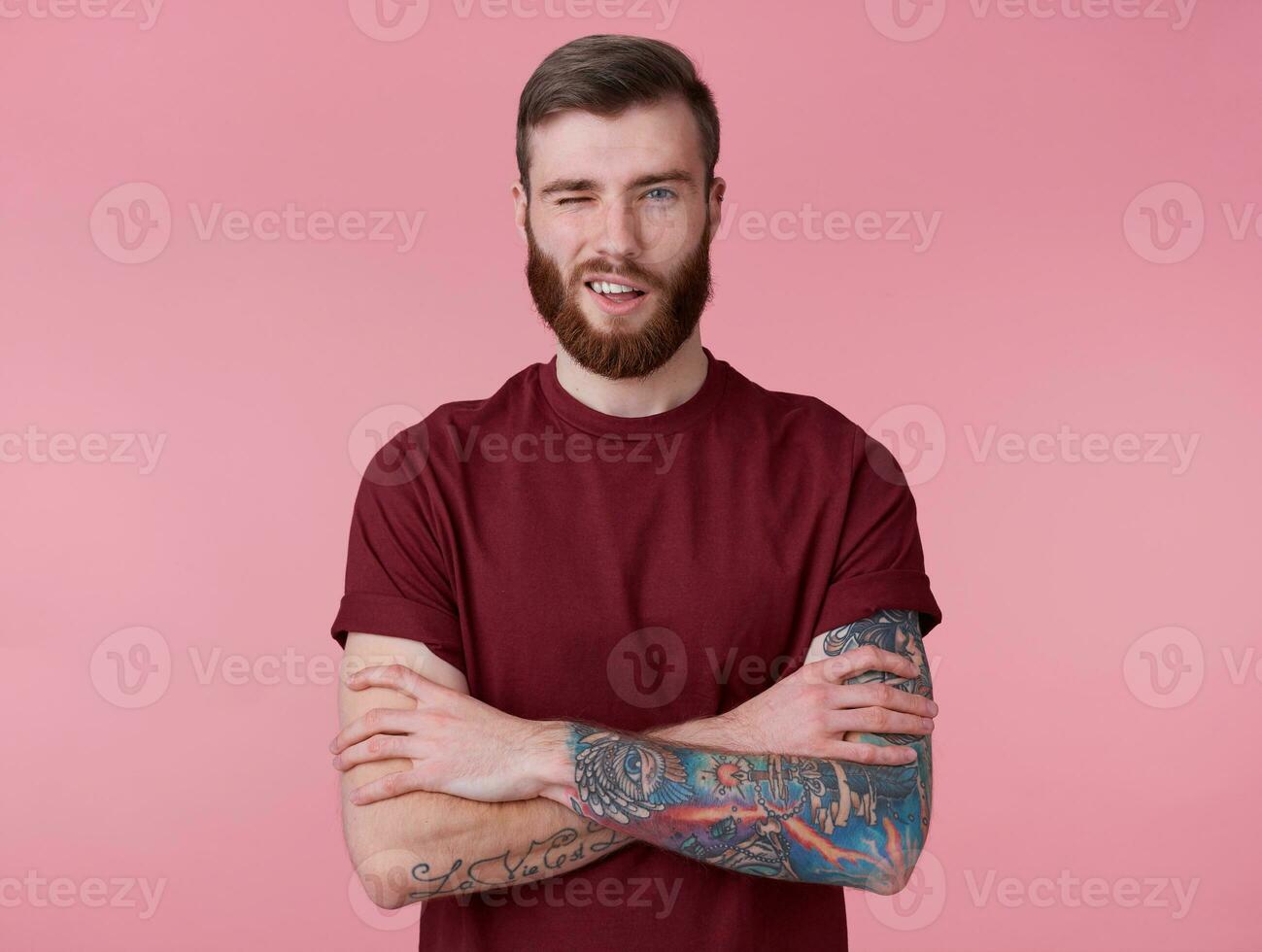 Young handsome positive red bearded guy in blank t-shirt, looks at camera, wink and broadly smiles, stands over pink background and wink. photo