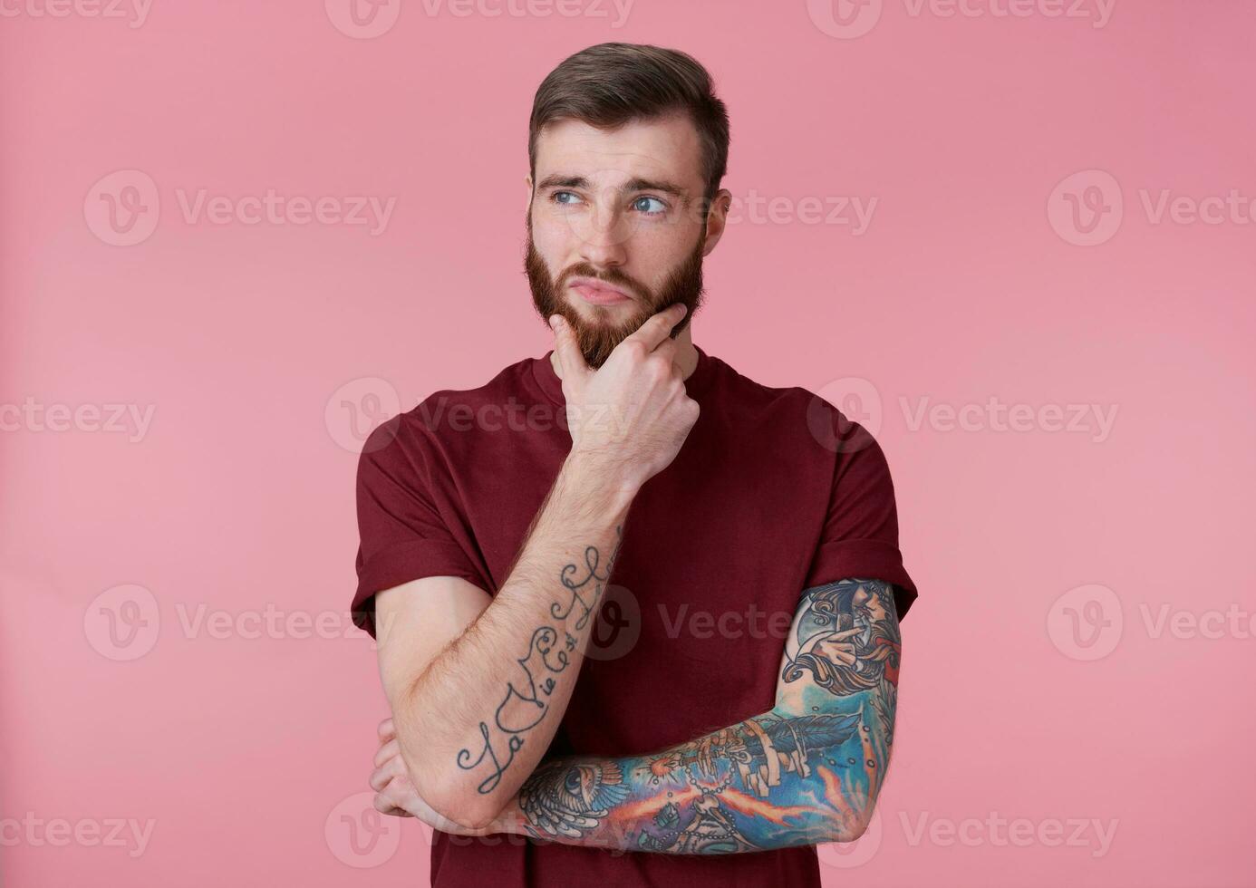 Portrait of young handsome thinking tattooed red bearded man in red t-shirt, looks away and touches the chin, stands over pink background. photo