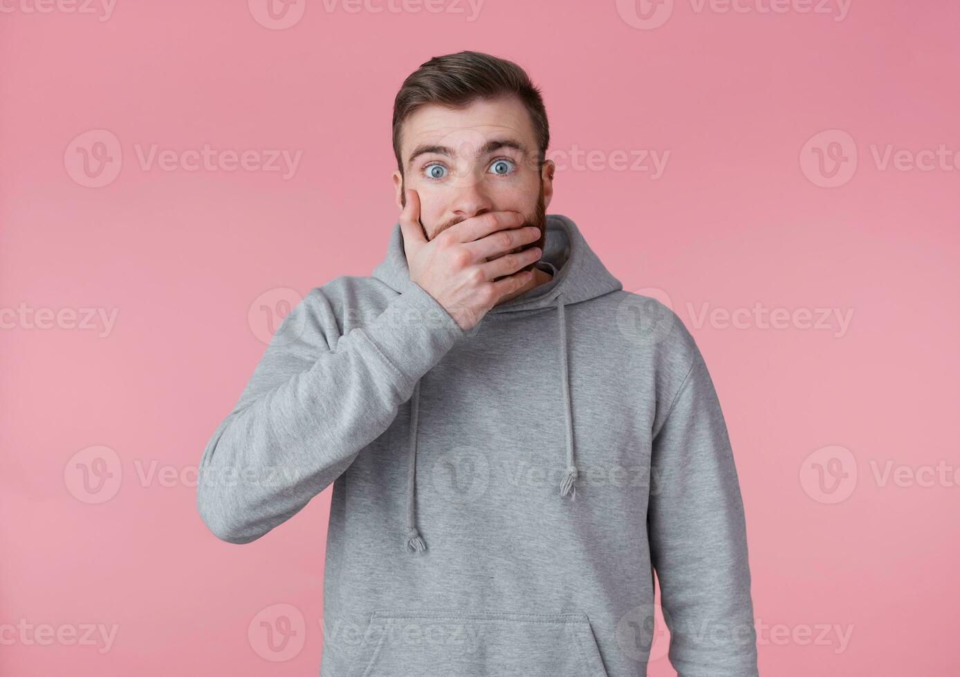 Portrait of young handsome shocked red bearded man in gray hoodie, hears unbelievable news, looks surprised and covers mouth with palm, stands over pink background with wide open eyes. photo