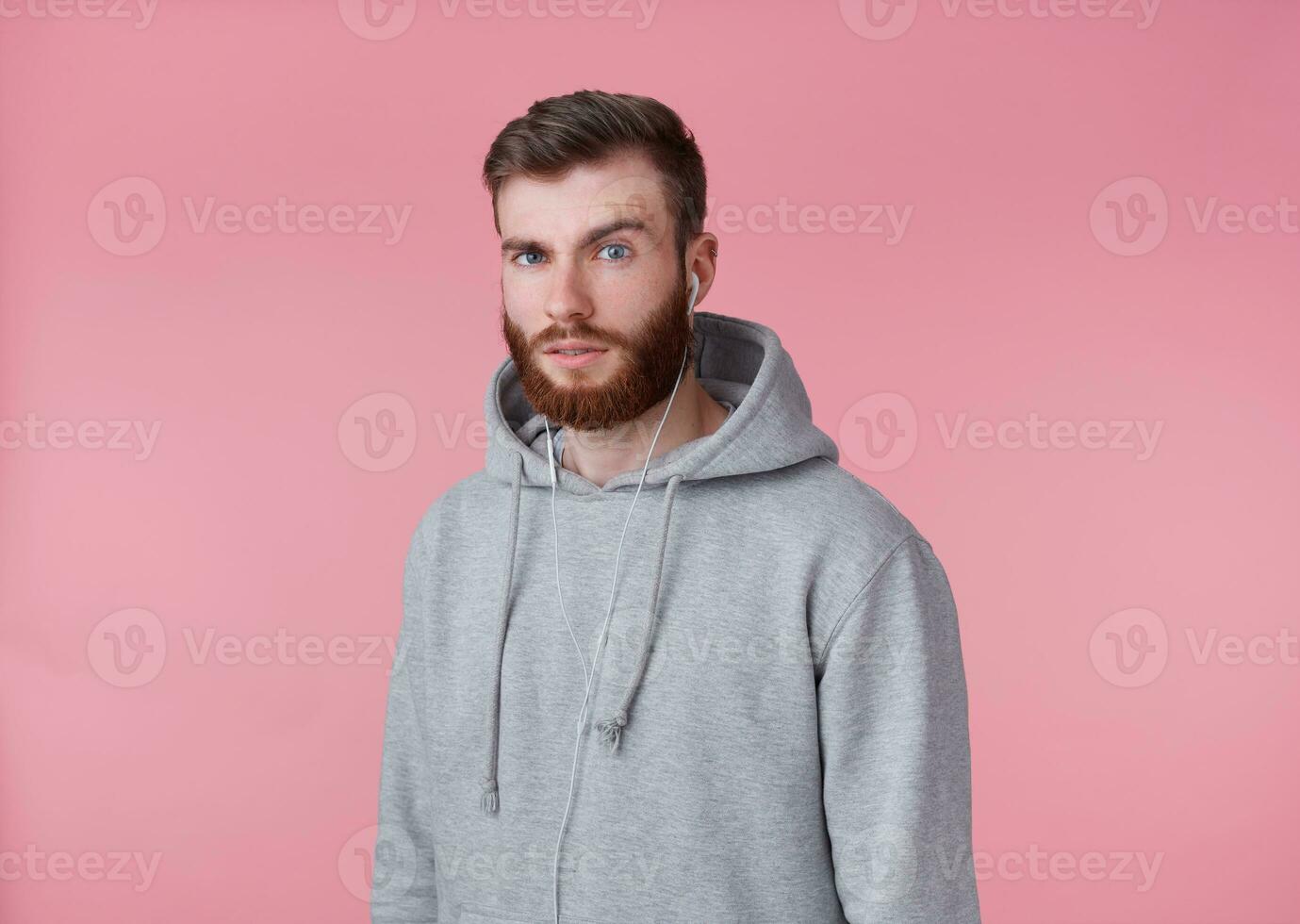 retrato de joven atractivo rojo barbado hombre en gris sudadera, mira bueno y calma, sonrisas, escuchando canción en auriculares, disfruta el música, soportes terminado rosado antecedentes. foto