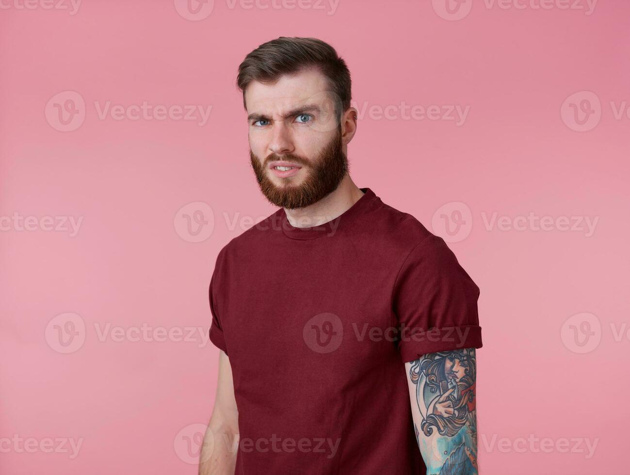 Photo of young handsome frowning misunderstanding red bearded man in blank t-shirt, stands over pink background looks at the camera with disgust.