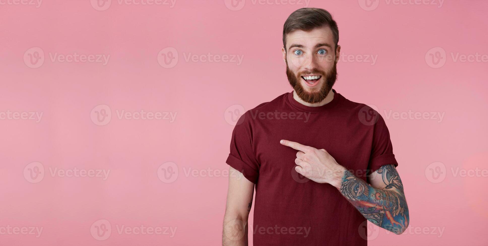 Portrait of happy amazed young handsome red bearded man in red shirt, wants to draw your attention to the left at copy space, pointing fingers and look at him, stands over pink background. photo