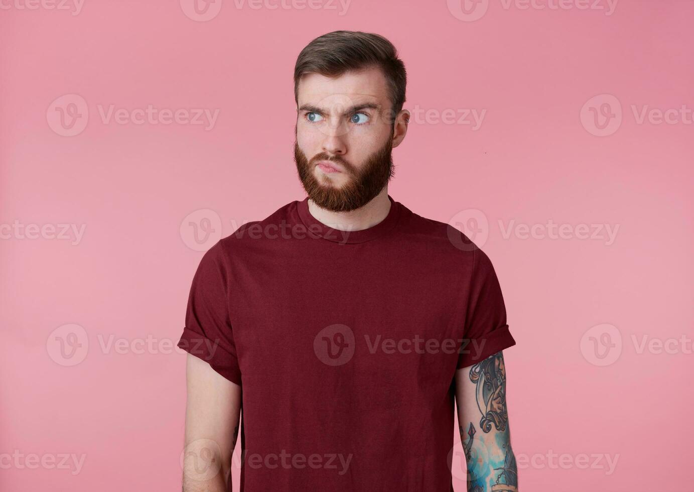 Portrait of young handsome tattooed misunderstanding red bearded man in red t-shirt, stands over pink background, thinking about something, looks away. photo