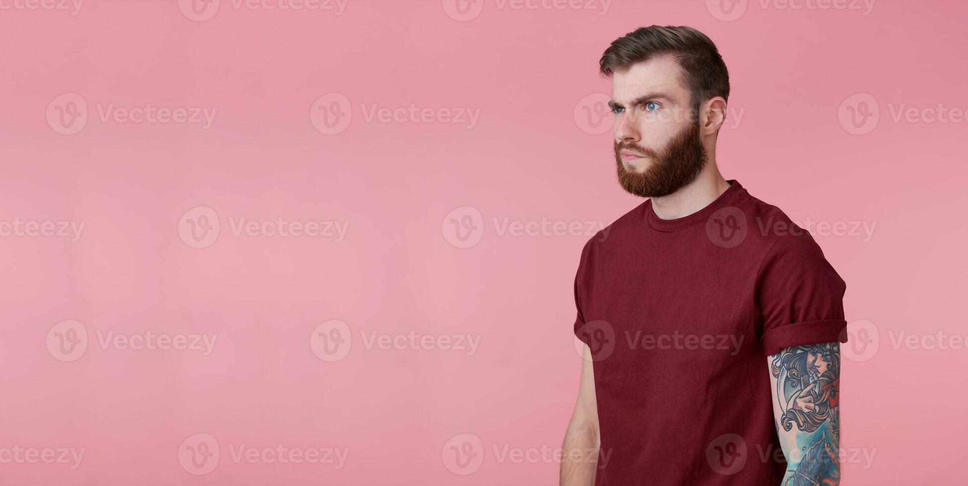 Portrait of young handsome misunderstanding red bearded man in red t-shirt, stands over pink background, looks to copy space on the left side, stands over pink background. photo