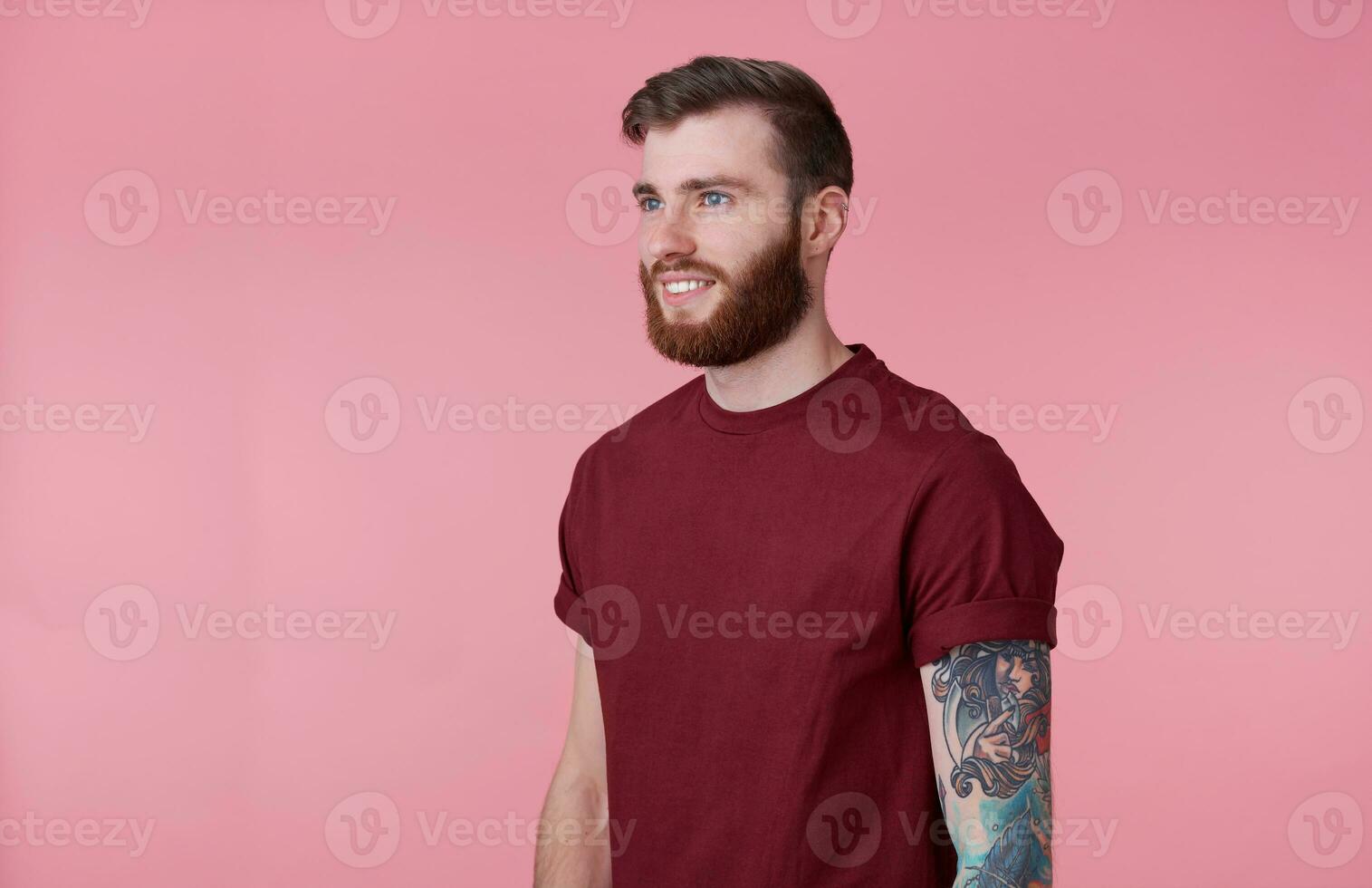 Photo of young handsome cheerful red bearded tattooed man in red t-shirt, looks happy and broadly smiles, looks away, stands over pink background.